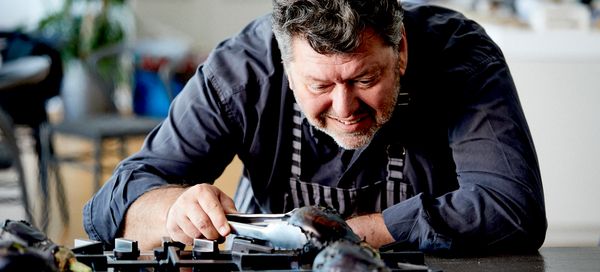 Ένας σεφ επιθεωρεί προσεκτικά ένα πιάτο με πολύχρωμα, περίτεχνα παρασκευασμένα φαγητά, επιδεικνύοντας την καλλιτεχνική του μαγειρική παρουσίαση.