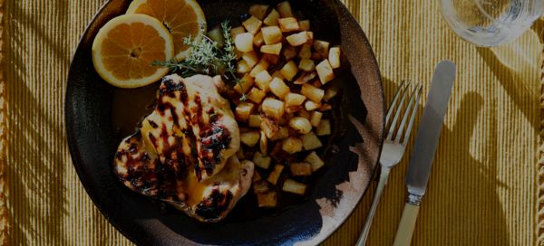 Ψητό στήθος κοτόπουλου με σημάδια από το γκριλ, σερβιρισμένο με χρυσές πατάτες σε κύβους και φέτες λεμονιού, σε σκούρο πιάτο.