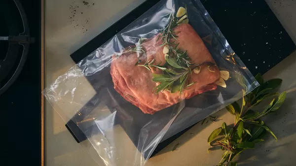 A wooden cutting board with a raw piece of meat and a knife, surrounded by tomatoes, garlic, onions, and fresh buns.