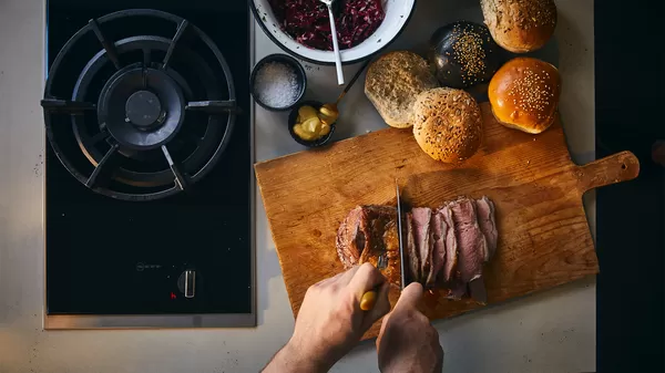 A person slices cooked meat on a wooden board, surrounded by burgers, condiments, and a stove, creating a meal scene.