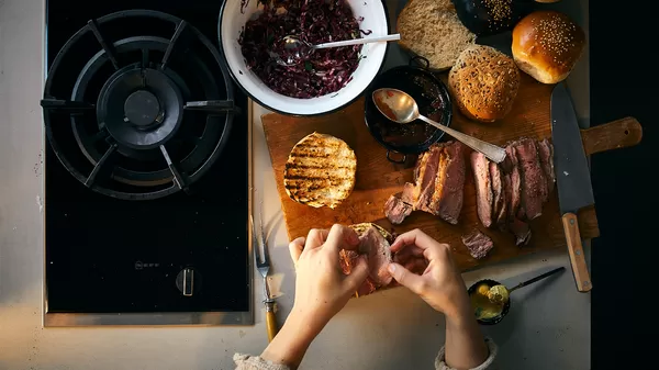 A person's hands assemble a meal with grilled beef, rolls, and red cabbage salad on a wooden board, beside a gas stove.