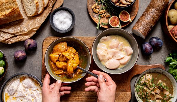 Mani che tengono una ciotola con bocconcini di pollo marinati accanto a una ciotola con pollo crudo affettato, circondate da pane, fichi e sale