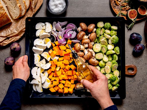 Vista dall&rsquo;alto di teglia con zucca, cavoletti, funghi, cipolla rossa e mozzarella mentre una mano aggiunge olio